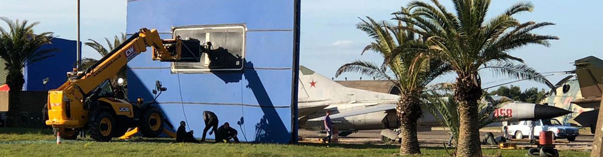 Telehandler holding a blue screen beside aircraft and palm trees on location